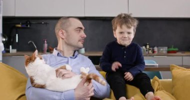 Sakin bir baba, üç gün sakallı, tüylü bir kedi ve heyecanlı bir çocukla kanepeye oturur. Duygusal çocuk, rahat stüdyo dairesinde şakacı bir tokat atmaya başladı.