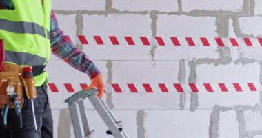 Mühendis alet çantasını büyük bir apartman dairesindeki merdivene yerleştirdi. Erkek müteahhit parlak uyarı bandına ve tuğla duvara karşı yürür ve yenileme çalışmalarına başlar