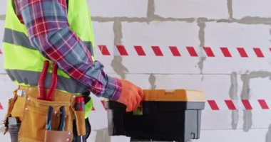 İnşaatçı alet çantasından tuğla duvara karşı şerit alıyor. Parlak renkli eldivenler ve özel yansıtıcı yelek giyen adam yenileme odasında çalışmaya hazırlanıyor.