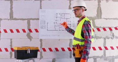 Uzman, kat planlarını analiz eder ve parmaklarıyla yerleşime dokunur. Parlak eldivenli inşaatçı yenileme sırasında koridorda duran alet çantası ve uyarı bandının önünde duruyor.