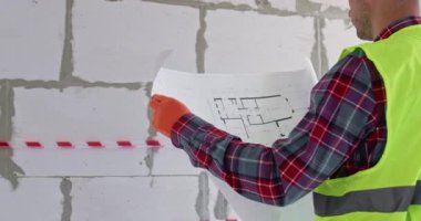 Parlak kask takan bina planıyla apartman planını karşılaştırıyor. Sakallı mühendis, büyük tuğla duvarın önünde duran yenileme işleminden önce planlama çalışması yapıyor.