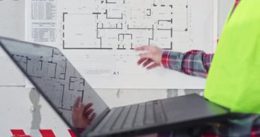 Adam dizüstü bilgisayarda çalışıyor. Sabit bina düzenine parmağıyla dokunuyor. Yansıtıcı yelek mühendisi elinde bilgisayarla, yenileme sırasında kat planıyla ilgili verileri analiz ediyor.