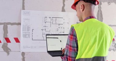 Mühendis apartman planlarını incelemek için modern teknolojiyi kullanıyor. Bilgisayarı elinde tutan inşaatçı geniş salondaki duvara yerleştirilmiş büyük kat planının önünde duruyor.