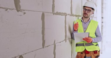 İnşaat uzmanı evdeki beton blokları incelerken not alıyor. Sert şapkalı erkek mühendis, özel materyal ve yaklaşan iş hacmini değerlendiriyor