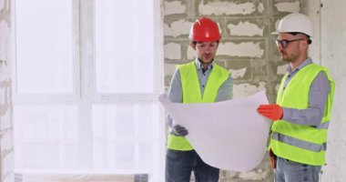 Erkek uzmanlar kat planının ellerde tutulacağını konuşuyorlar. Miğfer ve güvenlik yelekleri kullanan yapıcılar, konuşma sırasında büyük bir daireyi inceler ve çalışma sürecini kapsar