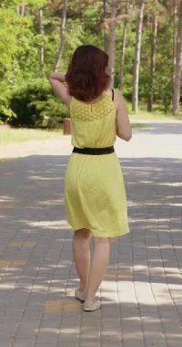 Şık bir kadın ağaçlarla kaplı park yolunda güvenle yürür. Sarı yazlık elbiseli kahverengi saçlı kadın parkta huzurlu bir şekilde yürümekten hoşlanıyor. Dikey çekim