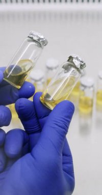 Lastik eldivenli laboratuar teknisyeni örneklerin rengini ve berraklığını karşılaştırıyor. Laboratuvar çalışanı, laboratuvar ışığı altında, farklılıkları tespit etmek için enjeksiyon ilacı şişelerini inceliyor.