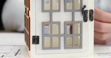 Adam masasında oturup banknotları dikkatle sayıyor. Minyatür ev modelinde indirim tabelası var. Mimar parayı sayar ve toplamı istenen fiyatla karşılaştırır