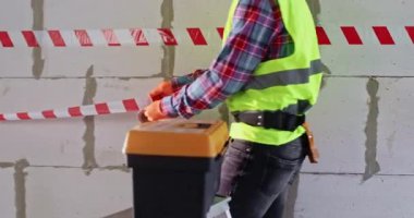 Uzman, yenileme bölgesini işaretlemek için parlak çizgili şerit kullanıyor. Yansıtıcı yelekli adam tadilata hazırlanıyor. Elinde profesyonel aletlerle büyük bir valizin yanından geçiyor.