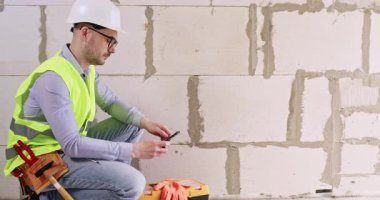 Güvenlik yeleği giyen ustabaşı, duvar yapımını analiz etmek için akıllı telefon kullanıyor. Sert şapkalı adam işi hızlandırmak ve bilgi almak için modern teknolojiyi kullanıyor.