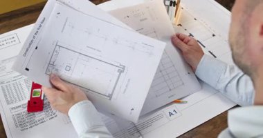 Tecrübeli bir mimar masada oturur ve farklı dairelerin çizimlerini inceler. Gömlekli bir ofis çalışanı yatak odasının planına çok dikkat eder.