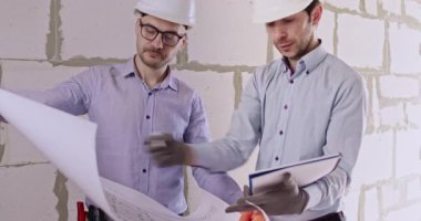 Uzmanlar kat planlarını inceler ve tartışmalar sırasında not alırlar. Sert şapkalı mühendisler apartman planlarını inceler ve duvara karşı duran kağıtlara bilgi yazarlar.