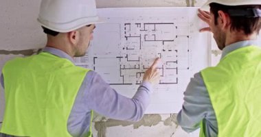 Uzmanlar, duvarına apartman planı asılmış kağıt çarşafları açıyorlar. Erkek ustalar gelecek çalışmaların aşamalarını cüruf briketlerinin yanında duran büyük görsel şema kullanarak analiz ederler