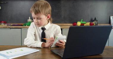 Tişörtlü ve kravatlı çocuk küçük not defterine ve kağıt çarşafa dizüstü kalemle ahşap masada yazıyor. İfade ve duruş göreve odaklanmayı iletir