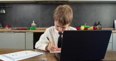 Kravat takan genç bir çocuk dizüstü bilgisayarının yanında açık kalemle deftere yazıyor. Çalışmaya odaklanmış bakışlar, çalışkanlığı ve öğrenme faaliyetlerine bağlılığı ortaya koyuyor