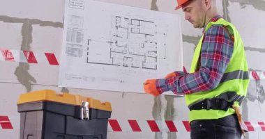 Miğfer konusunda uzman, geniş kat planındaki oda genişliğini ölçer. Usta, yaklaşan işin hacmini tahmin ediyor. Duvarın önündeki alet çantasının yanında duruyor.