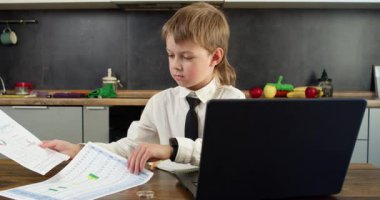 Kravatlı çocuk mutfaktaki masada açık dizüstü bilgisayarda kağıtları ve yazıları dikkatle inceliyor. Ciddi ifade, ödev ve projeyle ilgili göreve odaklanılmasını öneriyor