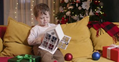 Cute boy takes shiny Christmas baubles out of Christmas house. Preschooler boy sits happily on comfortable couch and removes shiny decorations from toy house