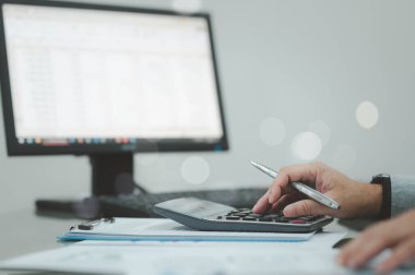 Bir kişi arka planda bilgisayar monitörü olan bir masanın üzerinde hesap makinesi kullanıyor. Kişi hesap makinesine odaklanmış ve bir görev üzerinde çalışıyor. Konsantrasyon ve verimlilik kavramı
