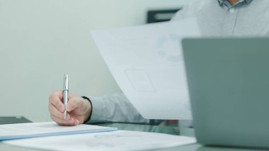 İşadamı bir kağıt parçasına kalemle yazıyor. Kağıt, bir dizüstü bilgisayarın yanındaki masada. İşadamı yazısına odaklanmış durumda ve sahne bir konsantrasyon ve verimlilik hissi veriyor.