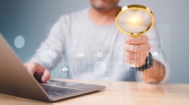 Bir adam dizüstü bilgisayarın üzerinde büyüteç tutuyor. Dizüstü bilgisayar ekranında bir saat, gülen yüz ve bir dolar işareti de dahil olmak üzere çeşitli simgeler ve semboller var.