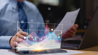 Borsa yöneticisi ekranda bir grafikle bir tablete yazı yazıyor. Grafik renkli ve bir sürü çizgisi var. Adam tablete yazmak için bir kalem kullanıyor.