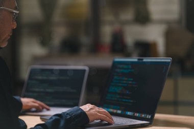 Programcı bir dizüstü bilgisayarda yazıp kodluyor. O gözlük takıyor ve klavyede bir eli var. Laptop açık ve ekran siyah. Adam işine odaklanmış.