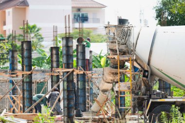 Bir çimento karıştırıcısı binaya beton döküyor. İşçiler sert kasklar ve güvenlik donanımı takıyor. İnşaat alanında çalışan insanlar yüzünden olay yeri çok yoğun ve hareketli.