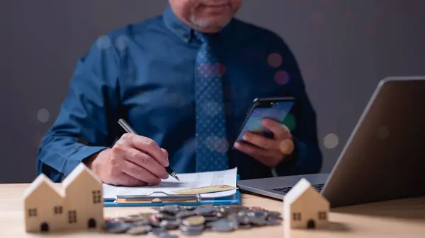 Bir iş adamı dizüstü bilgisayarı ve kalemi olan bir masada oturuyor. O, bir parça kağıda bir şey yazıyor. Masanın üzerinde madeni paralar da var. Finans, kredi, plan.