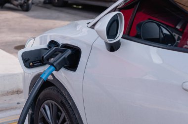 Sürdürülebilirlik kavramları. Elektrikli araçlar. Şarj istasyonunda beyaz bir araba şarj ediliyor. Araba bir otoparka park edilmiş.