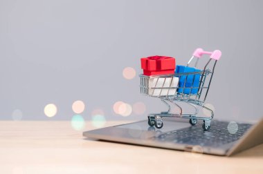 Renkli hediye kutularıyla dolu minyatür bir alışveriş arabası bir dizüstü bilgisayarın üzerinde oturur ve festival sezonlarında online alışverişin kolaylığını sembolize eder.