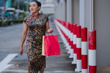 Stylish woman wears traditional cheongsam while walking down street, gracefully holding shopping bag amidst vibrant red and white bollards clipart