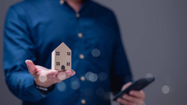 Mavi gömlekli kişi ahşap ev modelini elinde tutuyor. Modern ve şık ortamlarda akıllı telefon kullanırken ev sahibi olma yolculuğunu simgeliyor.