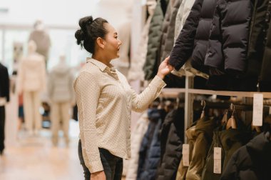 Kadın şık giyim mağazasında sıra sıra ceketleri karıştırıyor, bir sonraki moda alışverişini meşgul bir gülümsemeyle düşünüyor.
