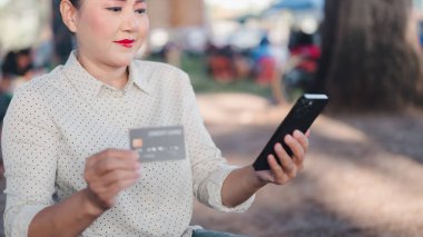 Bir kadın bankta oturuyor ve telefonuna bakıyor. Elinde bir kredi kartı tutuyor.