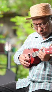 Hasır şapkalı bir adam ukulele çalıyor. Adam gözlük ve ekose gömlek giyiyor.