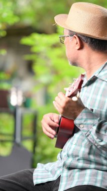 Hasır şapkalı ve gözlüklü bir adam ukulele tutuyor. O bir parkta bir bankta oturuyor.