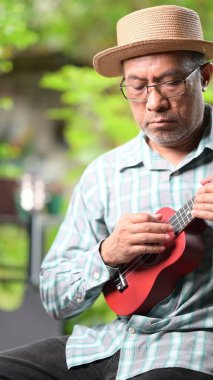 Hasır şapkalı bir adam ukulele çalıyor. Adam gözlük ve ekose gömlek giyiyor.