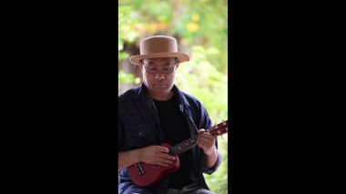 Bir adam parkta ukulele çalıyor. Hasır şapka ve gözlük takıyor.