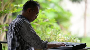 Bir adam klavye çalıyor. O gözlük takıyor. Klavye siyah. Adam bankta oturuyor.