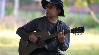 Parkta gitar çalan bir adam var. O bir şapka ve gözlük takıyor. Gitar siyah ve kayışı var.