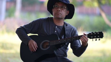 Şapkalı ve gözlüklü bir adam gitar çalıyor. O bir bankta oturuyor. Gitar siyah.