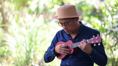 Hasır şapkalı bir adam küçük bir gitar çalıyor. Adam mavi bir gömlek ve gözlük takıyor.