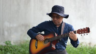 Mavi gömlekli ve şapkalı bir adam gitar çalıyor. Rahatlama ve rahatlama kavramı, adam enstrümanını çalarken çimlerin üzerinde oturuyor.