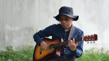 Mavi gömlekli ve şapkalı bir adam gitar çalıyor. Rahatlama ve rahatlama kavramı, adam enstrümanını çalarken çimlerin üzerinde oturuyor.