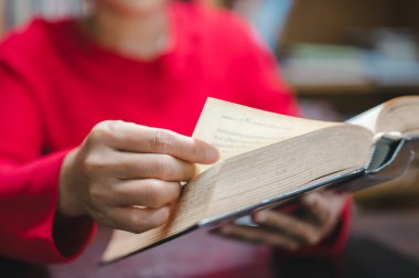 Canlı kırmızı süveter giyen bir kişi açık bir kitap tutar, sayfayı nazikçe çevirir, sıcak ve davetkar bir ortamda okuma ve öğrenme sevincini somutlaştırır.