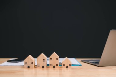 Resim, dizüstü bilgisayar ve belgelerin yanına dizilmiş, gayrimenkul, mimari ve planlama temalarını vurgulayan bir dizi ahşap ev modelini içeriyor.