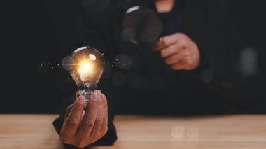 Parlayan ampulü elinde tutan kişi, yaratıcılığı ve yeniliği sembolize ederken, aynı zamanda parlak fikirlerin arayışında yeni perspektifleri incelemek için büyüteç kullanıyor.