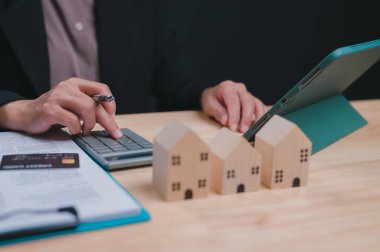 Kadın elinde hesap makinesi ve bir yığın kağıtla masada oturuyor. Evlerle ilgili bir proje üzerinde çalışıyor.