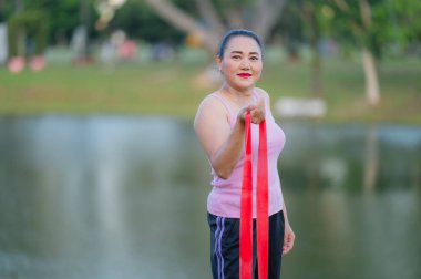 Ortaçağ Asyalı Kadın Park 'ta Kızıl Direniş Bandosu, Pembe Tank Üstü, Kırmızı Ruj, Doğal Makyaj, Yeşillik ve Suyun Ortasında, Kameraya Gülümseyen, Doğal Yaşam Tarzı Kucaklayan